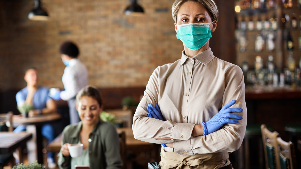 Waitress standing with arms crossed while wearing protective face mask and working in a cafe during coronavirus epidemic.