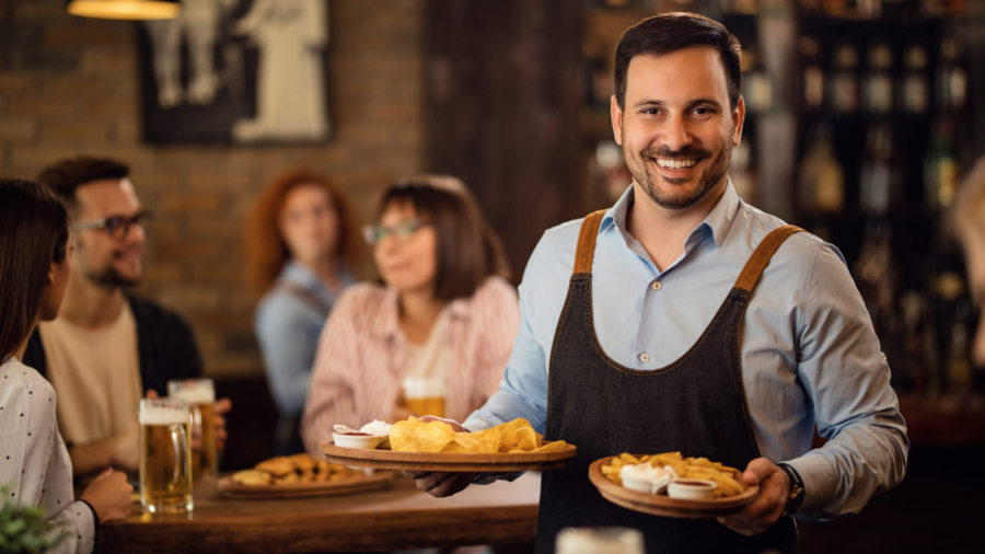 Happy waiter holding plates with food and looking at camera while serving guests in a restaurant.